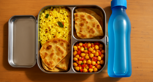 Open three-compartment stainless steel tiffin box with lemon rice, aloo paratha, and masala corn on a wooden desk